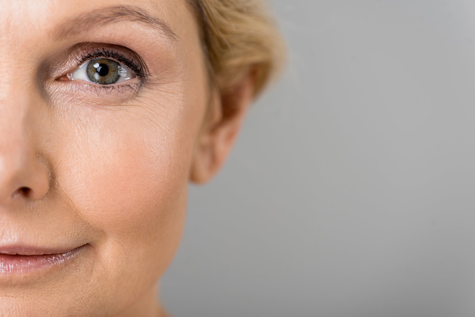 Close-up of a smiling middle-aged woman&rsquo;s face, fine lines visible around a bright eye against a soft grey background &mdash; the modern pursuit of youthful, healthy aging.