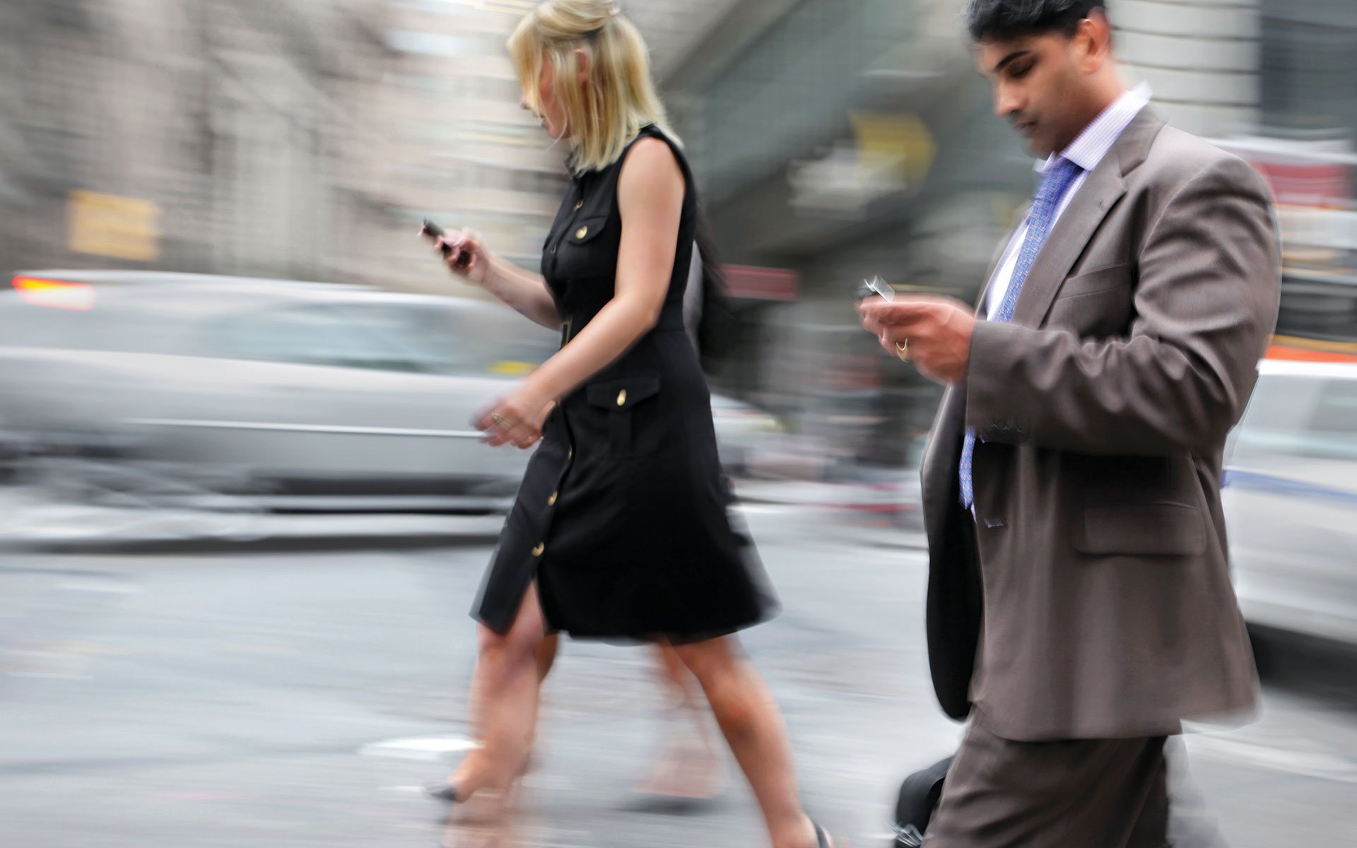 Commuters striding through a blurred city street, absorbed in their phones — the accelerating pace of modern life.