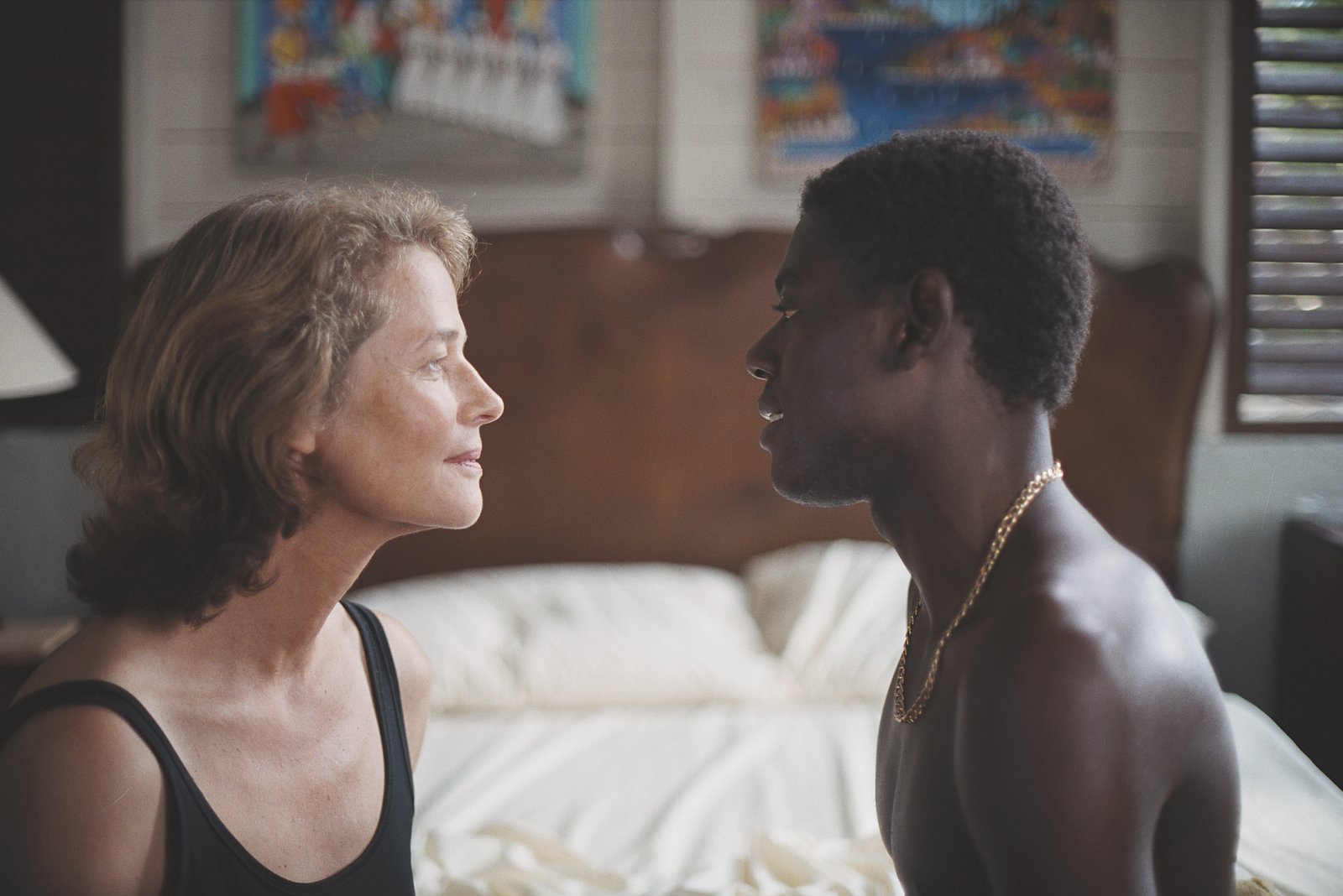 Film still from the 2005 movie Heading South: an older woman in a black tank top and a young shirtless man wearing a gold chain face each other closely in a sunlit Caribbean bedroom.